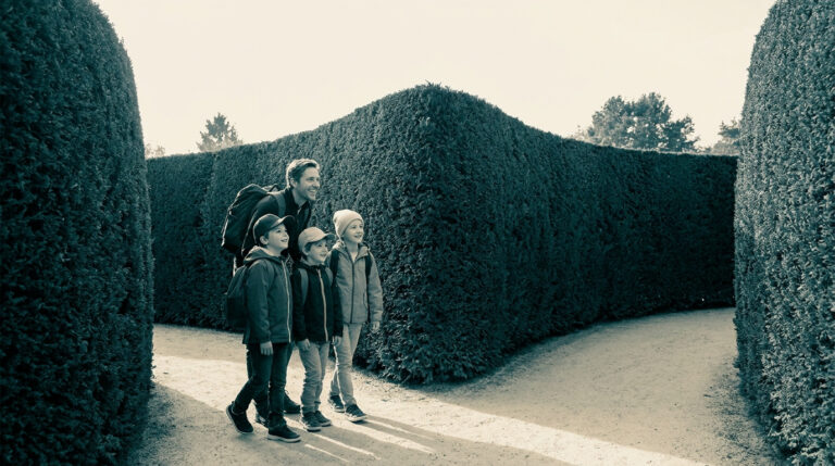 Un père et ses trois jeunes enfants explorent ensemble les allées d'un labyrinthe de haies hautes en extérieur.