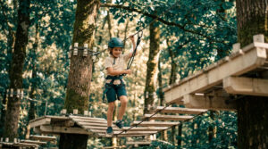 Un jeune garçon souriant traverse un pont de singe en accrobranche, équipé d'un casque et d'un harnais de sécurité.