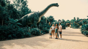Une famille observe une grande statue de brachiosaure dans un parc à thème entouré de végétation luxuriante.