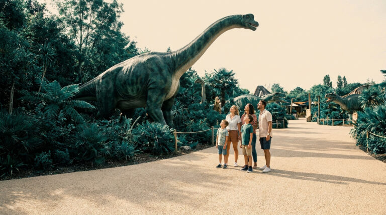 Une famille observe une grande statue de brachiosaure dans un parc à thème entouré de végétation luxuriante.