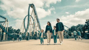 Une famille souriante marche dans les allées d'un parc d'attractions avec des montagnes russes en arrière-plan.