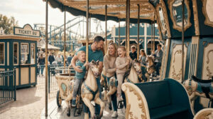 Une famille souriante s'amuse sur un manège à chevaux rétro dans un parc d'attractions avec une billetterie en arrière-plan.